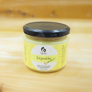 Op de afbeelding: Een glazen pot met een zwart deksel met een gele kaars erin. Het etiket op de pot staat "Ops Wax" en "Citronella" in het Grieks. Het etiket vermeldt ook "100% Natuurlijke Bijenwas Citronella Kaars" in het Engels.