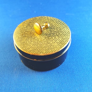 Puede incluir: Un gemelo de color dorado con una parte superior redonda y un pequeño botón redondo en el lateral. El gemelo está sobre una caja con un patrón negro y verde.