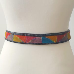 Op de afbeelding: Een bruine leren riem met een kleurrijk patchwork ontwerp. Het patchwork is gemaakt van verschillende gekleurde suède stukken die in een geometrisch patroon aan elkaar zijn genaaid.