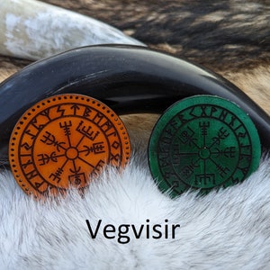 May include: Two leather Vegvisir compass charms, one orange and one green, with Norse runes around the edges. The charms are on a white furry surface.