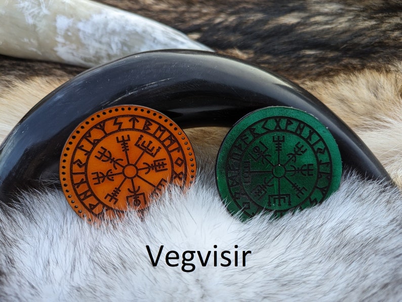 May include: Two leather Vegvisir compass charms, one orange and one green, with Norse runes around the edges. The charms are on a white furry surface.