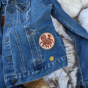 May include: A blue denim jacket with a brown leather patch featuring a stylized design of a horse. The patch is sewn onto the left pocket of the jacket.