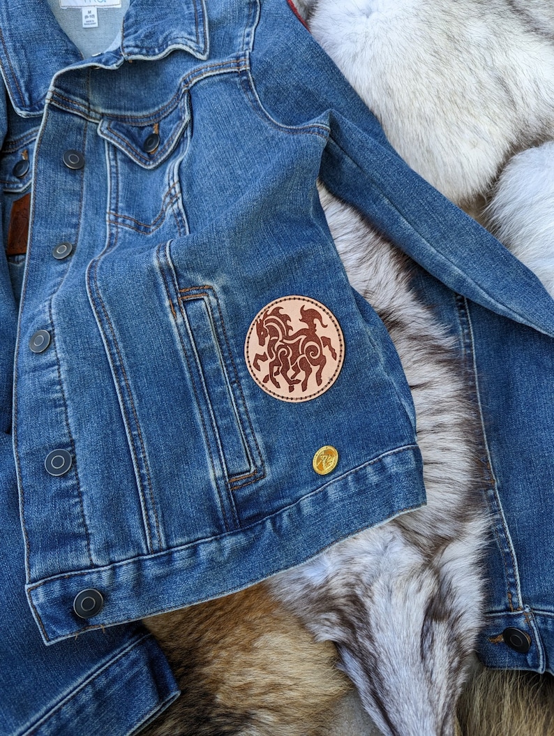 May include: A blue denim jacket with a brown leather patch featuring a stylized design of a horse. The patch is sewn onto the left pocket of the jacket.