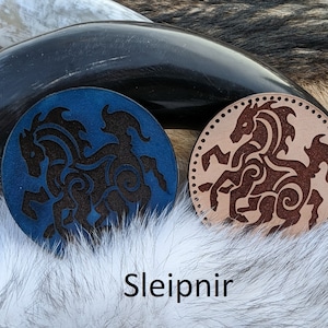 May include: Two leather coasters with a black silhouette of a horse with intricate designs. The coaster on the left is blue and the coaster on the right is brown. The text "Sleipnir" is printed below the coasters.
