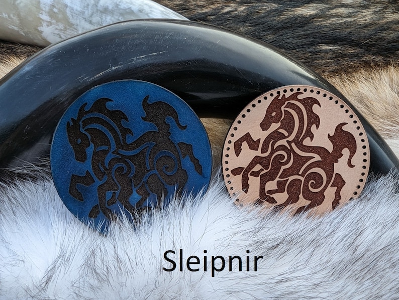 May include: Two leather coasters with a black silhouette of a horse with intricate designs. The coaster on the left is blue and the coaster on the right is brown. The text "Sleipnir" is printed below the coasters.