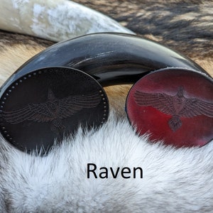 May include: Two leather coasters with a raven design. One coaster is black and the other is red.