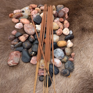 Peut inclure: Un paquet de bâtonnets d'encens bruns disposés sur un lit de pierres colorées. Les pierres sont de formes et de tailles variées, et certaines ont une finition lisse et polie.