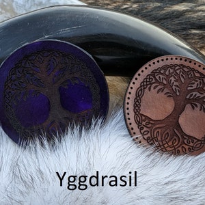 May include: Two leather coasters with a tree of life design. One coaster is brown with a light brown tree and the other is purple with a dark brown tree. The coasters are on a white furry surface. The text "Yggdrasil" is written below the coasters.