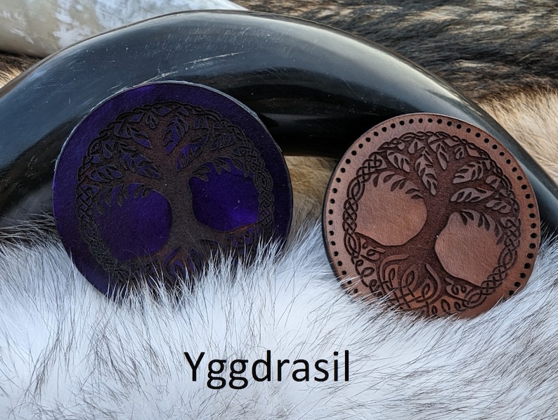 May include: Two leather coasters with a tree of life design. One coaster is brown with a light brown tree and the other is purple with a dark brown tree. The coasters are on a white furry surface. The text "Yggdrasil" is written below the coasters.
