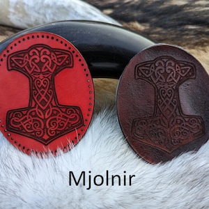 May include: Two leather patches with a Mjolnir design. One patch is red and the other is brown. The patches are on a white furry surface. The text "Mjolnir" is below the patches.