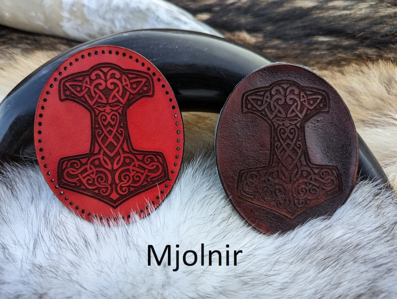 May include: Two leather patches with a Mjolnir design. One patch is red and the other is brown. The patches are on a white furry surface. The text "Mjolnir" is below the patches.