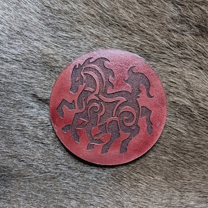 May include: A round, red leather coaster with a black silhouette of two intertwined horses. The horses are stylized with intricate details and a Celtic knot design.