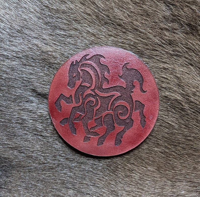 May include: A round, red leather coaster with a black silhouette of two intertwined horses. The horses are stylized with intricate details and a Celtic knot design.
