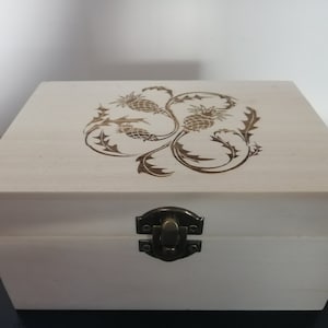 May include: A white wooden box with a hinged lid and a brass latch. The top of the box is decorated with a laser-etched design of stylized thistles.