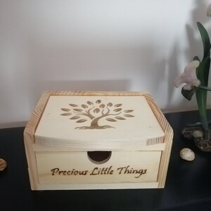 Puede incluir: Una caja de madera con un cajón. La parte superior de la caja tiene un diseño de árbol y el cajón tiene las palabras "Precious Little Things" grabadas.