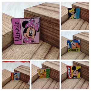 May include: Six colorful square pins featuring Disney characters. The pins feature Minnie Mouse, Tinkerbell, Goofy, Donald Duck, Pluto, and Mickey Mouse. Each pin has a character's name printed on it.