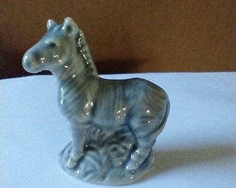May include: A small, gray ceramic figurine of a zebra standing on a base. The zebra has black stripes and is in a standing position.