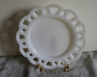 May include: A white, decorative plate with a scalloped edge and openwork design. The plate is round with a smooth center and is displayed on a gold-colored stand. The plate's design is reminiscent of vintage or antique dinnerware.