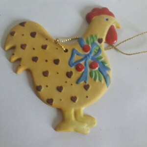 May include: A yellow ceramic rooster ornament with brown hearts, a blue bow, and red berries. The rooster has a red comb and a gold string for hanging.