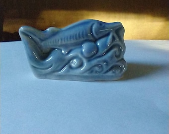May include: A blue ceramic figurine of a fish swimming in waves. The fish has a detailed design and is a decorative piece.