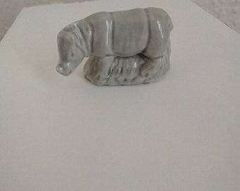 May include: A small gray ceramic figurine of a rhinoceros standing on a base of grass.