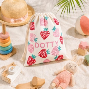 May include: A selection of children's toys, including a straw hat with a pink ribbon, a drawstring bag with strawberry print and the word "DOTTY", a wooden dog pull toy, and a crochet bunny.
