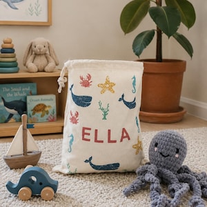 May include: A cream drawstring bag decorated with sea creatures, including whales, seahorses, and starfish, with the name "ELLA". A wooden sailboat, a blue wooden car, and a grey crocheted octopus are in the foreground. The bag is filled.