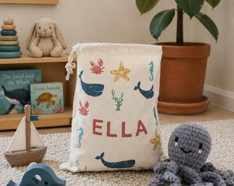 May include: A cream-colored drawstring bag with a nautical theme, featuring blue whales, seahorses, starfish, and the name "ELLA" in red. A wooden sailboat, a blue wooden car, and a gray crocheted octopus are in the foreground.