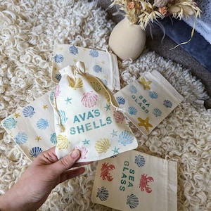 May include: Four small, white fabric drawstring bags with different designs. The bags are decorated with blue, yellow, and red sea-themed illustrations. The text on the bags reads "Sea Glass", "Ava's Shells", and "Sea Glass".