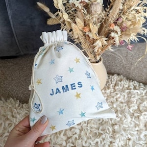 May include: A white drawstring bag with a blue, yellow and green star pattern. The bag has the name "JAMES" printed in blue on the front.