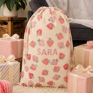 May include: A beige drawstring gift bag adorned with a strawberry pattern in shades of red and pink. The name "SARA" is printed in pink. Gift-wrapped presents with ribbons are arranged around the bag.