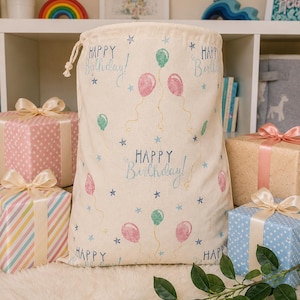 May include: A beige drawstring gift bag printed with "Happy Birthday!", balloons, and stars. The bag is surrounded by wrapped presents with ribbons and bows, creating a celebratory atmosphere.