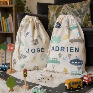 May include: Two cream drawstring bags with space and transport illustrations. One bag has the name "JOSÉ" and the other "ADRIEN" in navy blue. Toy cars, a plane, and a road are in the foreground, creating a playful scene.