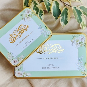 May include: Two green and gold gift boxes with floral designs and the text "Eid Mubarak" in Arabic calligraphy. The boxes also include the text "Love, The Zia Family".