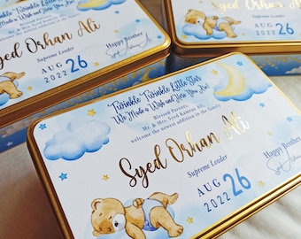 May include: A blue and gold gift box with a teddy bear sleeping on a cloud. The box has a gold trim and the text "Twinkle Twinkle Little Star We Made a Wish and Here You Are!" and "Syed Orhan Ali Supreme Leader AUG 26 2022".