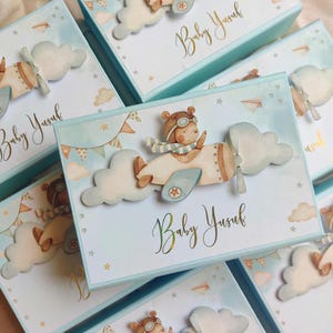 Puede incluir: Varias cajas de regalo azul claro con un diseño de acuarela que presenta un oso en un biplano, nubes y estrellas. Las cajas tienen el nombre "Baby Yusuf" en escritura dorada. Decoradas con recortes 3D.