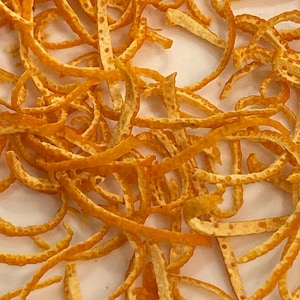May include: Close-up of orange zest, a pile of thin, dried orange peels, with a light orange color and a slightly rough texture.
