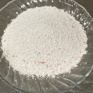 May include: A clear glass bowl filled with a mound of white powder with small, light brown particles. The bowl is round and sits on a dark surface. The powder appears to be a fine, granular substance.