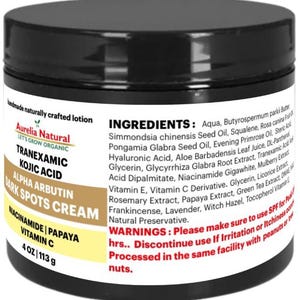 May include: A white jar with a black label that reads "Aurelia Natural Let's Grow Organic Tranexamic Kojic Acid Alpha Arbutin Dark Spots Cream Niacinamide Papaya Vitamin C 4 oz/113 g". The label also lists the ingredients and warnings for the cream.
