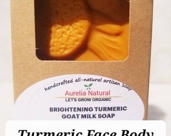 Op de afbeelding: Een oranje kurkuma geitenmelk zeep in een bruine kartonnen doos met een venster. De zeep heeft de tekst "Turmeric Face Body Soap". Het label zegt "Brightening Turmeric Goat Milk Soap".