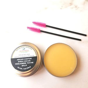 Puede incluir: Un bote de bálsamo facial metálico con la tapa quitada, que revela un bálsamo amarillo. La etiqueta dice "Argan Castor Shea Cocoa Fenugreek Thyme Balm". Dos varitas de rímel rosas y negras están junto al bálsamo. El bálsamo pesa 28 g.