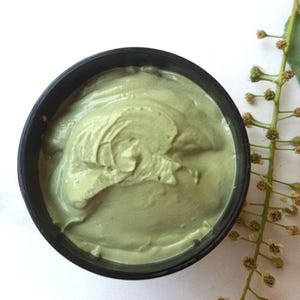 Puede incluir: Primer plano de un recipiente negro lleno de una sustancia cremosa de color verde claro. La crema tiene una textura espesa y arremolinada. Una ramita de una planta con pequeños capullos marrones está a la derecha. El fondo es blanco.