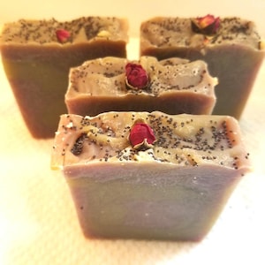 May include: Four rectangular bars of soap with a muted mauve and green color palette. Each bar is topped with a scattering of dark poppy seeds and a single dried red rosebud embedded into the textured surface of the soap.
