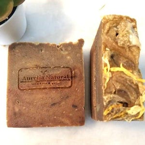 Könnte beinhalten: Zwei handgemachte Seifenstücke. Eines ist rechteckig mit dem geprägten Text "Aurelia Natural Let's Grow Organic". Das andere hat eine strukturierte Oberfläche und gelbe Akzente. Die Seifen sind braun.