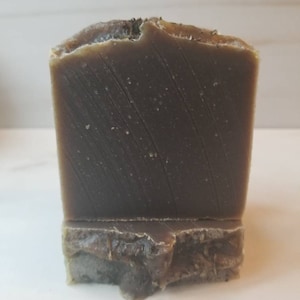 Puede incluir: Una barra de jabón marrón oscuro con una superficie rugosa y texturizada. El jabón está sobre una base del mismo material, pero de color marrón más claro.