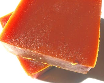 May include: Two square pieces of a reddish-orange food item, possibly a preserve or sweet, are stacked. The top piece is in focus, showing a smooth surface and a slightly rough edge. The bottom piece is partially visible.