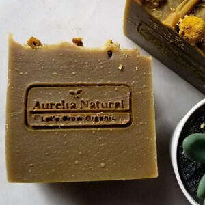 May include: A brown bar of soap with a label that reads "Aurelia Natural Let's Grow Organic". The soap has dried flowers and herbs embedded in it.