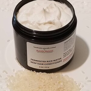 May include: A black jar filled with white cream, labeled "Fermented Rice Water Deep Hair Conditioner." The jar is 4 oz (113 g). A pile of white rice grains sits in front of the jar on a white plate. The product is from Aurelia Natural.