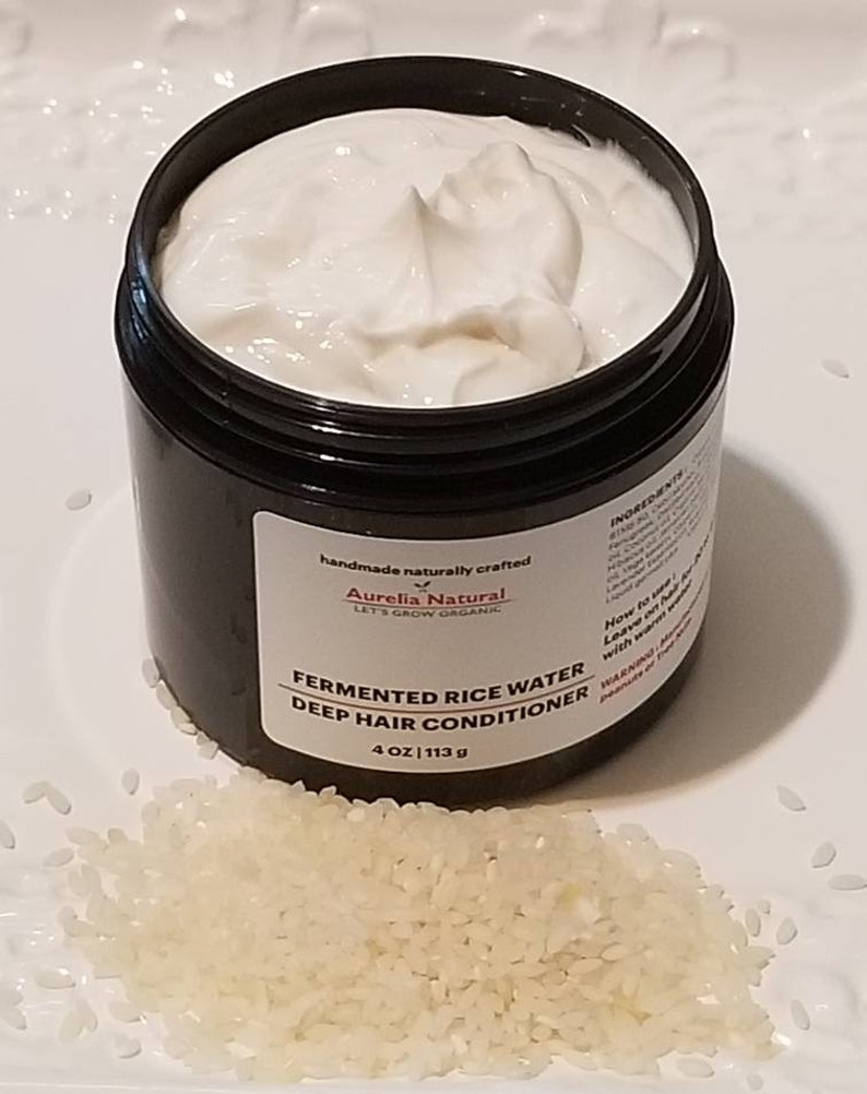 May include: A black jar filled with white cream, labeled "Fermented Rice Water Deep Hair Conditioner." The jar is 4 oz (113 g). A pile of white rice grains sits in front of the jar on a white plate. The product is from Aurelia Natural.