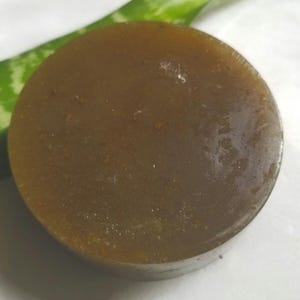 May include: A round, brown soap bar with a slightly translucent appearance. The soap has a smooth surface and appears to be handmade. A green and white aloe vera leaf is visible in the background.